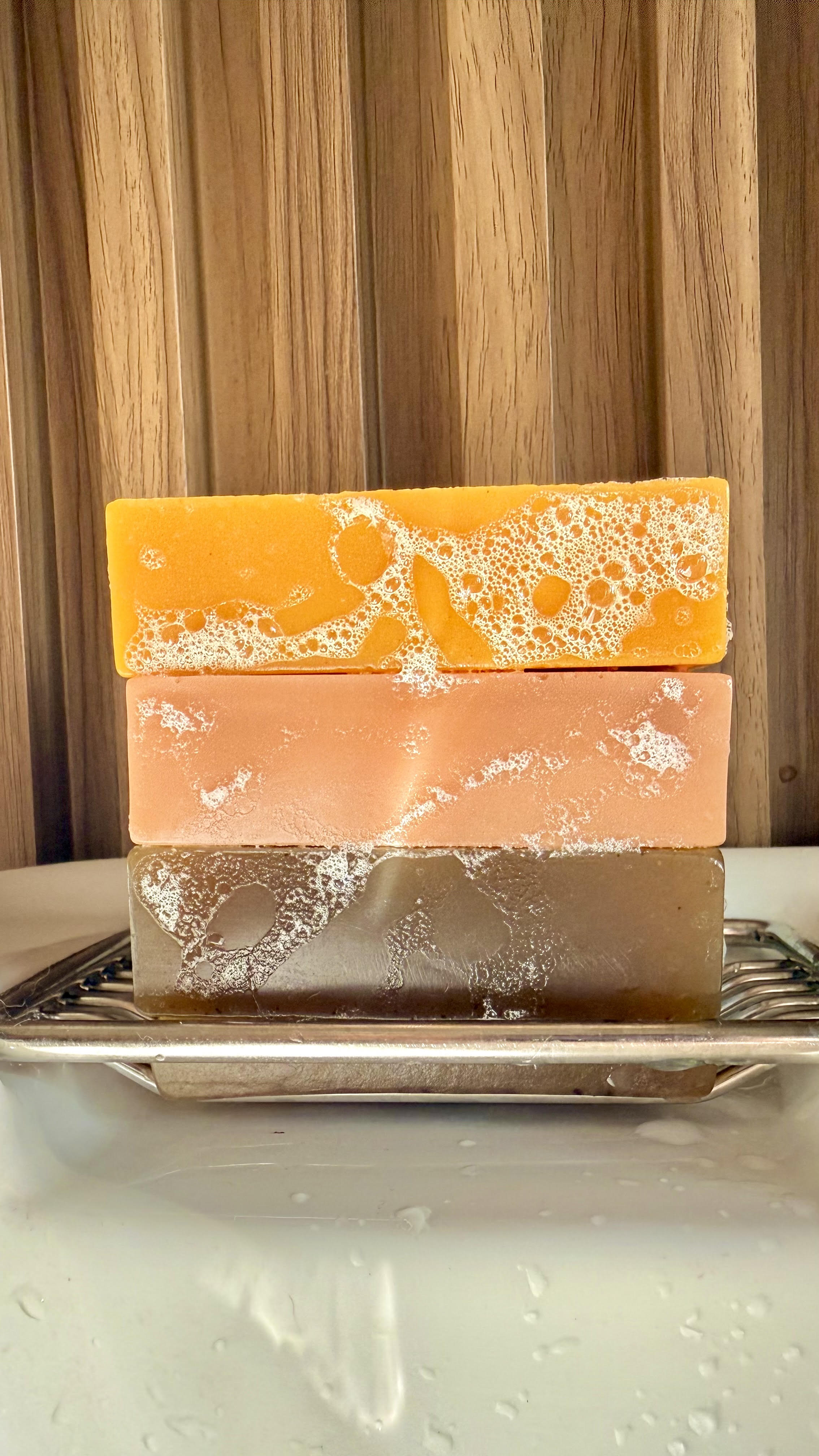 Three stacked soap bars with visible lather on a metal soap dish against a wooden background.